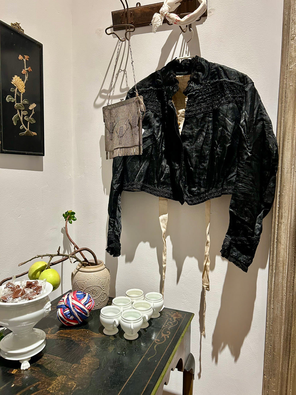 An antique black silk jacket and a beaded mesh bag hanging above a painted black table with white porcelain cups