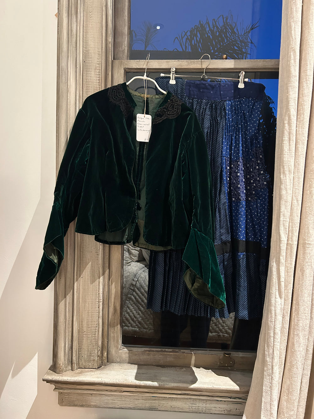 A 19th-century green velvet jacket and a patched indigo skirt hanging in a tall window at dusk